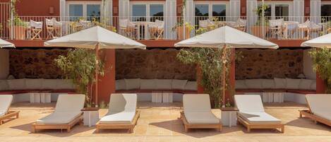 A poolside area with lounge chairs and umbrellas, a stone wall, and a building with balconies and windows in the background.