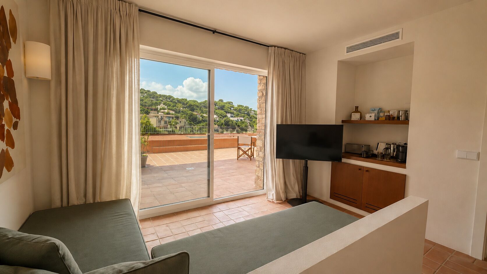 A hotel room with a sofa, a flat-screen TV, a kitchen area, and a sliding glass door leading to an outdoor patio.