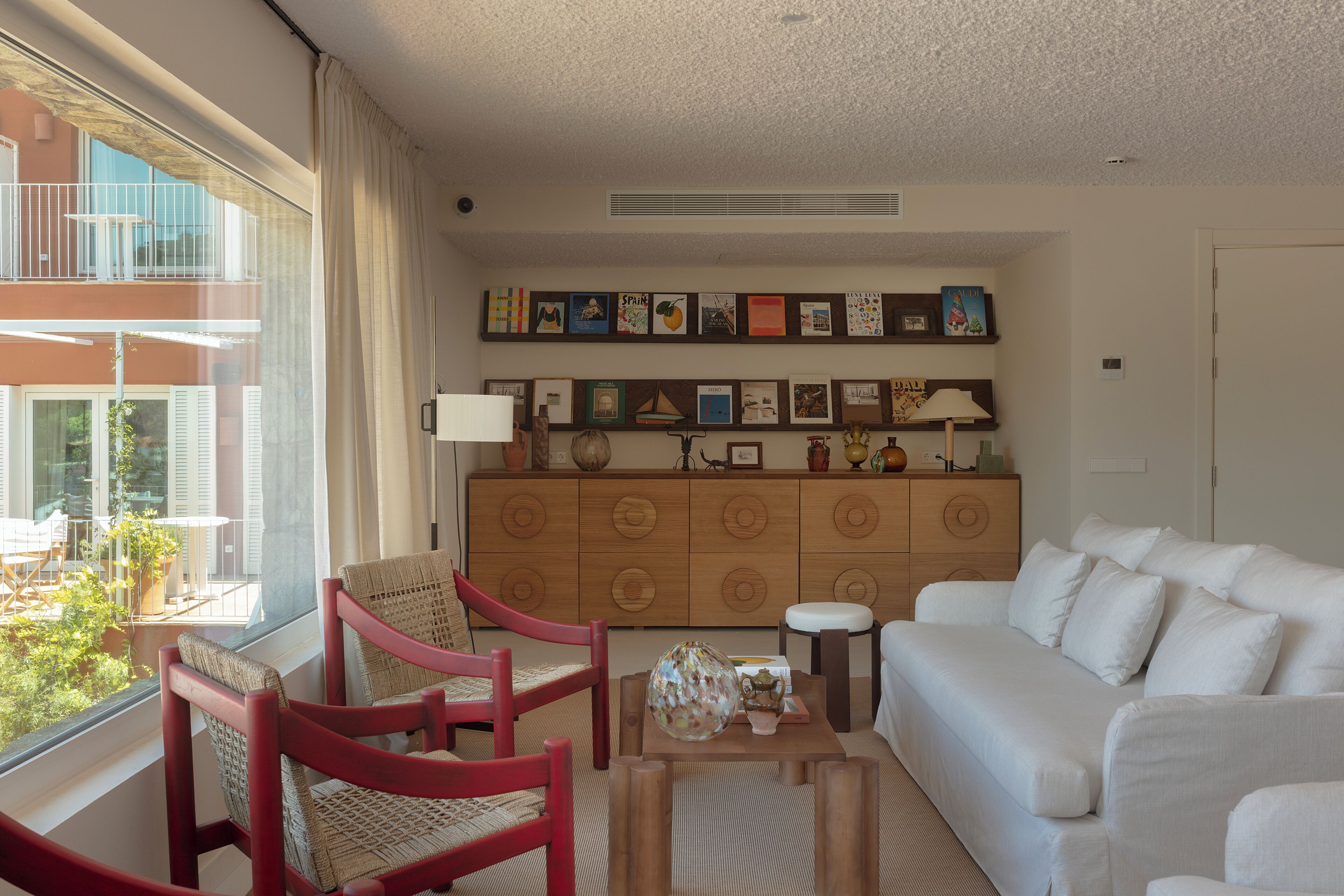 A living room with a white sofa, red chairs, a wooden cabinet with artwork, and a view of a balcony with outdoor furniture.