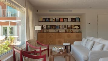 A living room with a white sofa, red chairs, a wooden cabinet with artwork, and a view of a balcony with outdoor furniture.