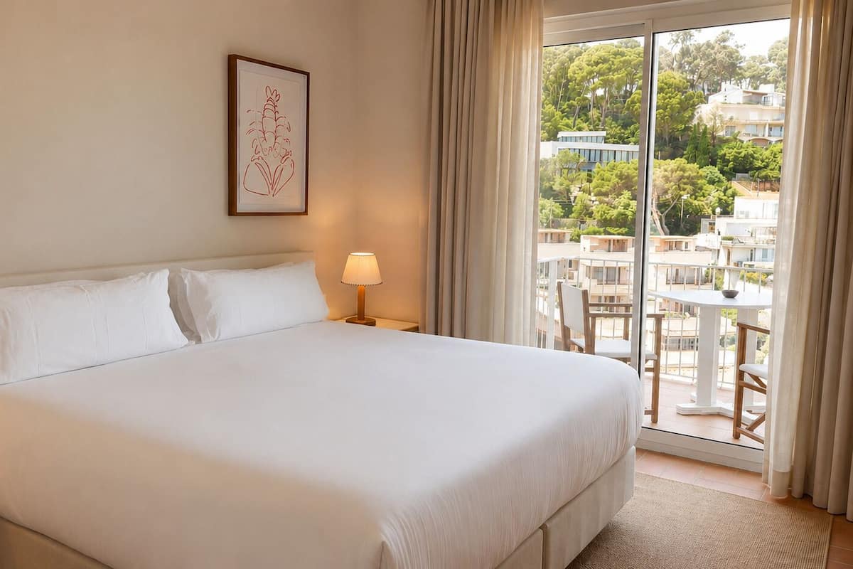 A hotel room with a large bed, a bedside table with a lamp, a framed picture on the wall, and a balcony with outdoor furniture.