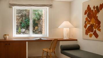 Habitació amb escriptori, cadira i banc, finestra amb vista a vegetació, i làmpada penjada a la paret.
