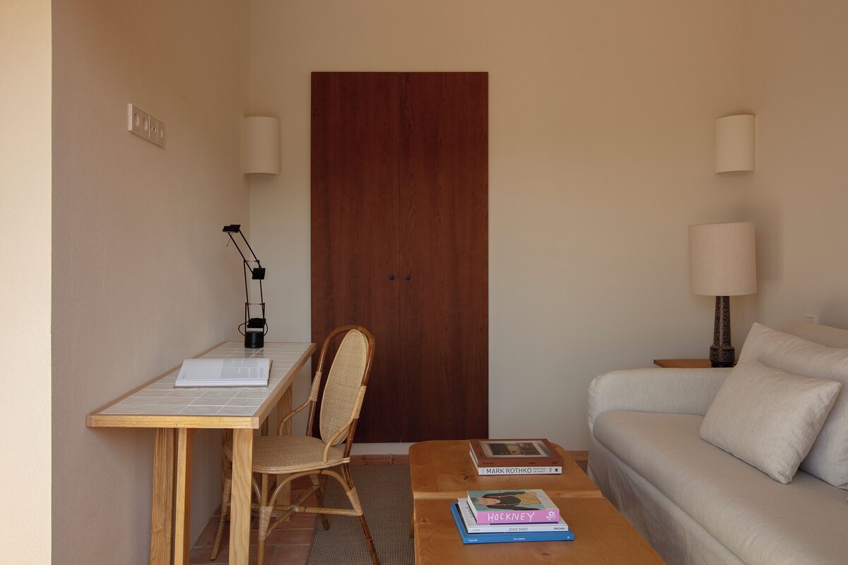 Une pièce avec un bureau en bois, une chaise en osier, une petite table avec des livres, un canapé et une porte en bois.
