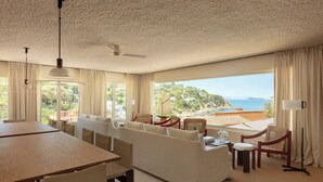Ein großzügiger Wohnbereich mit einem großen Fenster, das Meer und Hügel zeigt, einem Esstisch und einer Couch mit Blick.