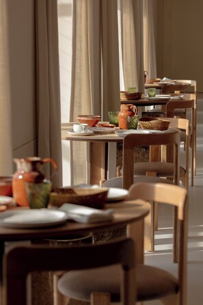 Un espace repas avec des tables et des chaises en bois, une fenêtre avec des rideaux, et divers plats et ustensiles disposés sur les tables.