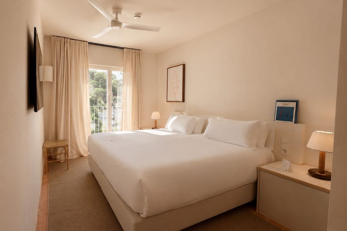 A hotel room with a large bed, a ceiling fan, a window with curtains, and a framed picture on the wall.