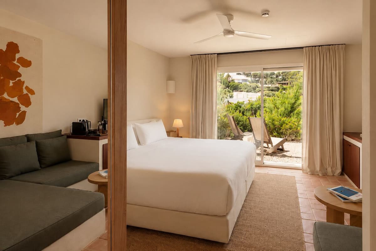 A hotel room with a bed, a sofa, a desk, a chair, a ceiling fan, and a view of greenery outside.