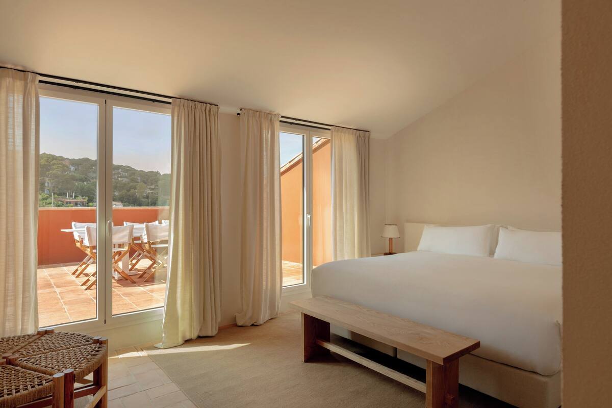 A hotel room with a large bed, a wooden bench, a wicker chair, and a balcony with a table and chairs.
