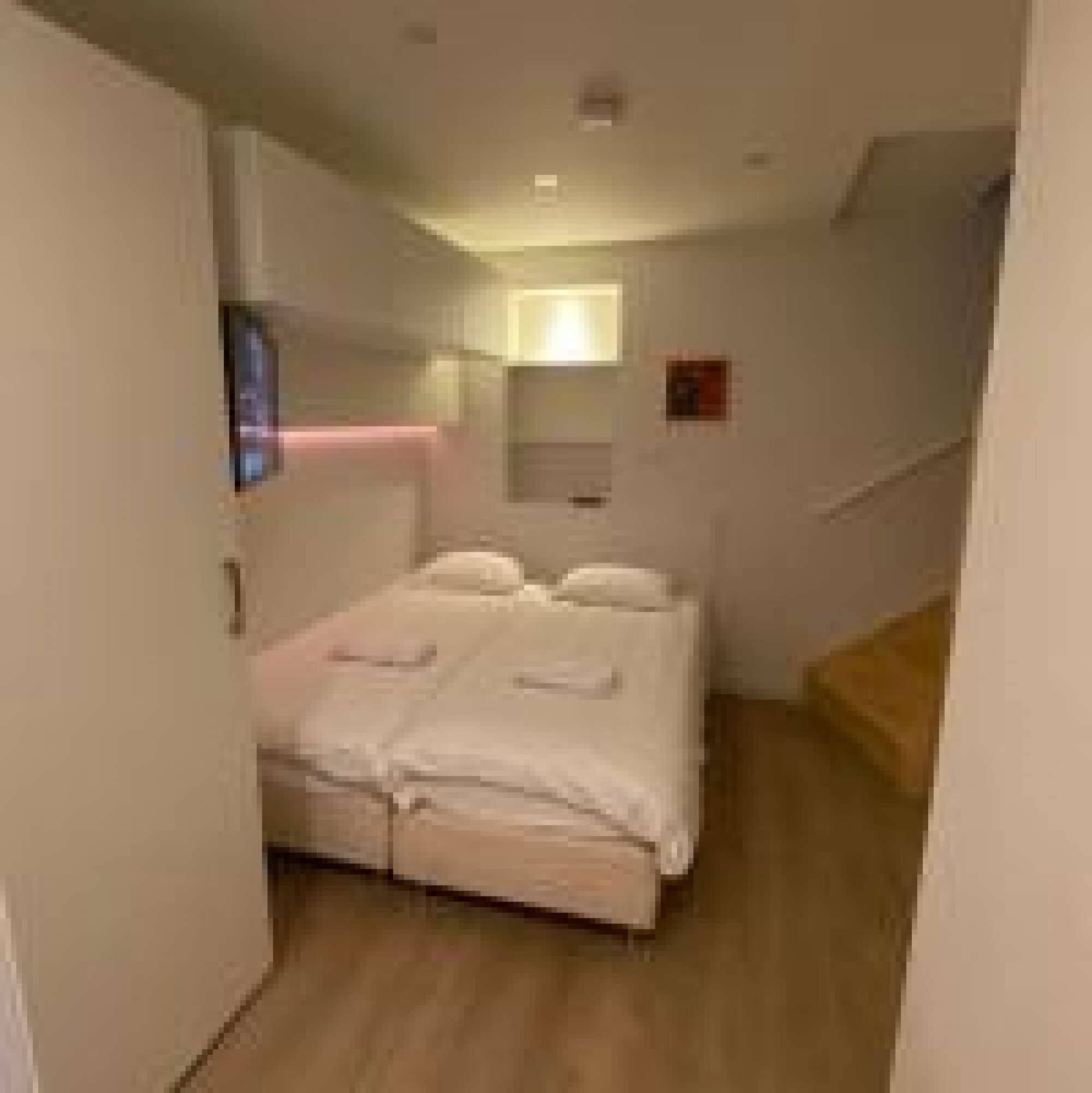 Une chambre à coucher avec un lit, une table de chevet, un téléviseur fixé au mur et une petite fenêtre.