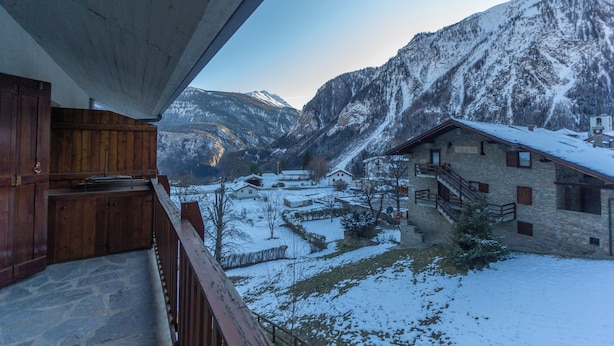Ein Holzbalkon mit Blick auf ein verschneites Dorf, mit Steinhäusern und Bergen im Hintergrund.