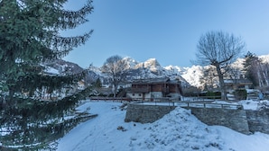 Schneebedeckte Landschaft mit einem Holzbau, einer Steinmauer und verschneiten Bergen im Hintergrund.