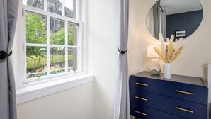 A room with a large window, a round mirror, a dark blue dresser with gold handles, and a vase with dried plants. - Castle Esplanade Apartment (Stirling)