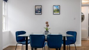 A dining area with a glass table, four blue chairs, a potted plant, and two framed cityscape pictures on the wall. - Castle Esplanade Apartment (Stirling)