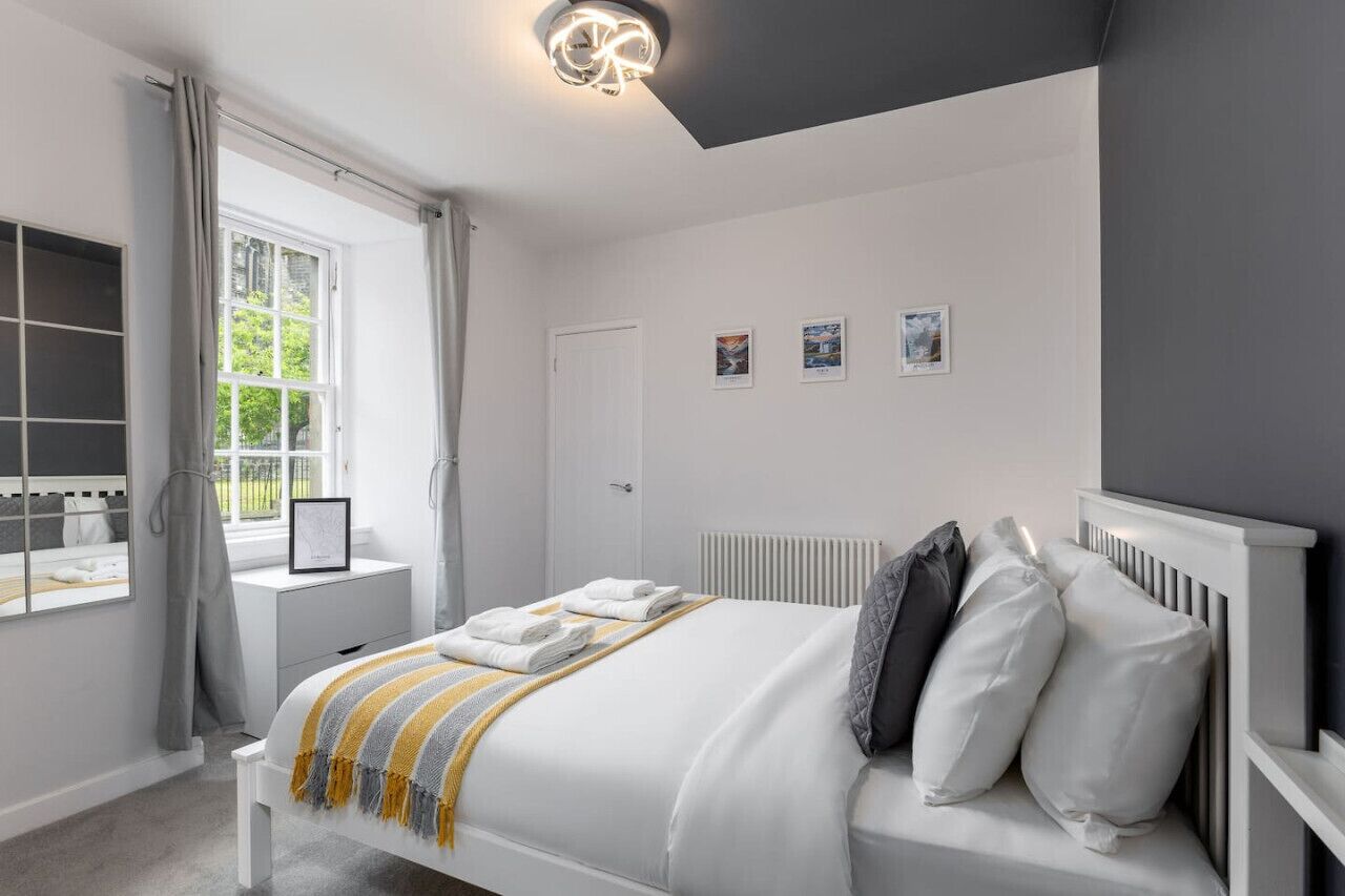A bedroom with a bed, a bedside table, a window with curtains, a radiator, and wall-mounted pictures.