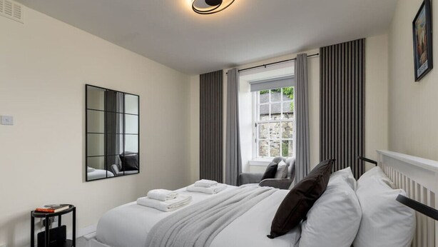 A modern bedroom with a bed, a bedside table, a mirror, and a window with curtains. - Castle Esplanade Apartment (Stirling)