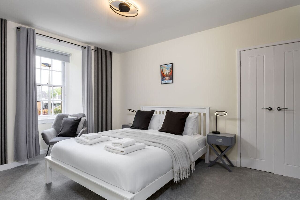 A modern bedroom with a white bed, grey curtains, a grey chair, a bedside table with a lamp, and a door.