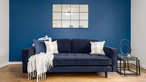 A dark blue sofa with white and plaid cushions, a white throw blanket, and a small black side table with a ring and a candle holder. - Castle Esplanade Apartment (Stirling)