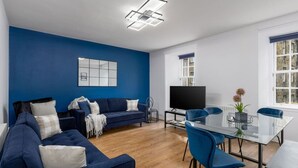 A modern living room with a blue accent wall, a large flat-screen TV, a glass dining table with blue chairs, and a cozy sofa set. - Castle Esplanade Apartment (Stirling)