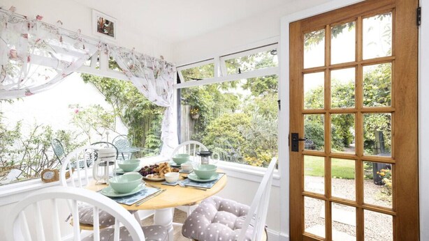 Un coin repas chaleureux avec une table en bois dressée pour deux personnes, entouré de chaises blanches ornées de coussins à pois. Une porte en bois avec des panneaux de verre donne accès à un jardin.