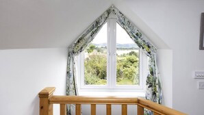 Une pièce dotée d’une grande fenêtre à cadre blanc et de rideaux à motifs floraux, d’une rampe d’escalier en bois, et offrant une vue sur la verdure et les collines.