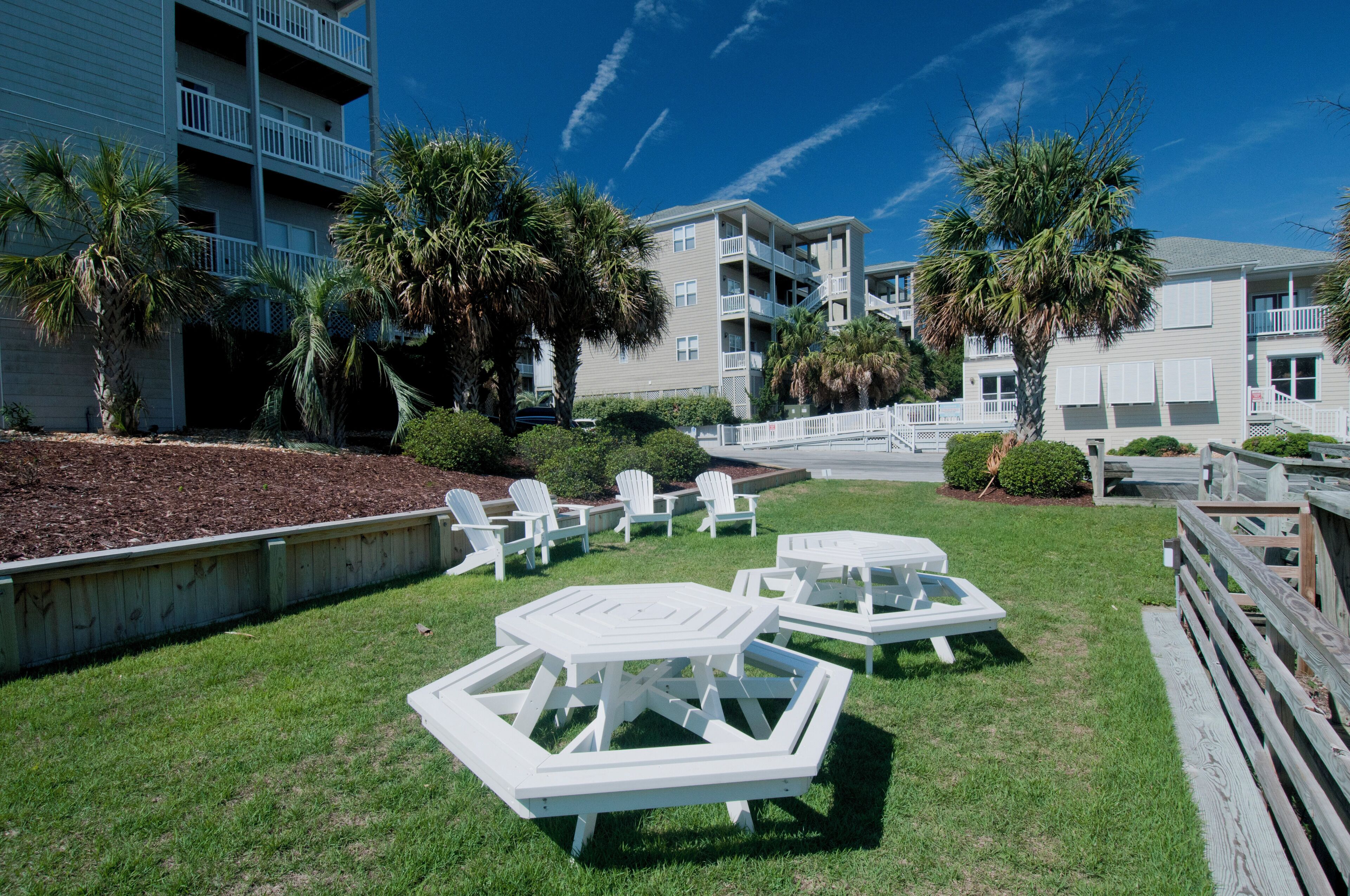 A grassy outdoor area with white tables and chairs, surrounded by buildings with balconies and palm trees.
