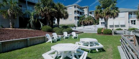 A grassy outdoor area with white tables and chairs, surrounded by buildings with balconies and palm trees.