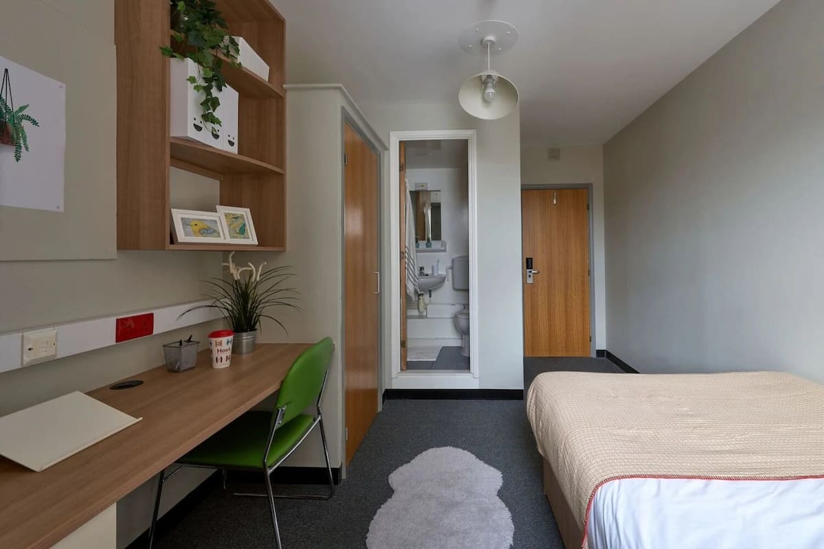 A hotel room with a bed, a desk with a chair, a small plant, and a bathroom visible through an open door.