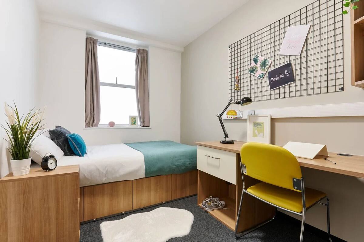A compact bedroom with a bed, bedside table, desk, chair, and a window with curtains.