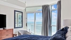 A hotel room with a large bed, a TV on a wooden dresser, a chair, and a balcony with a view of the ocean.