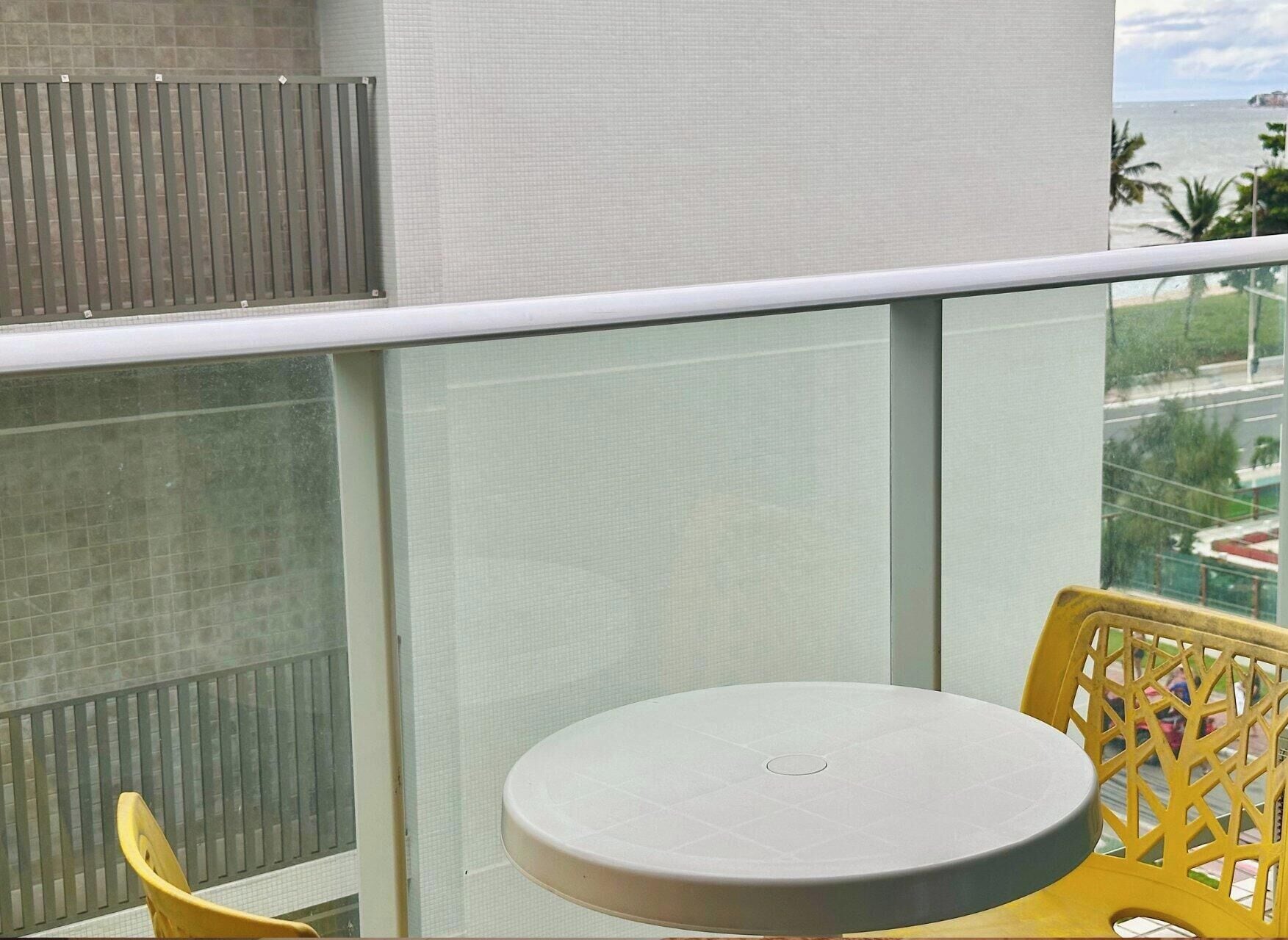 Varanda com uma mesa redonda e cadeiras amarelas, com vista para uma área costeira com edifícios e palmeiras.