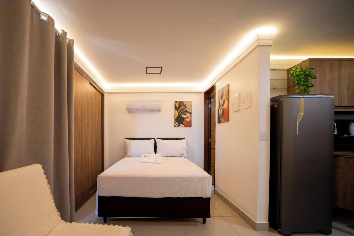 A hotel room with a bed, a sofa, a kitchen area, and an air conditioning unit.