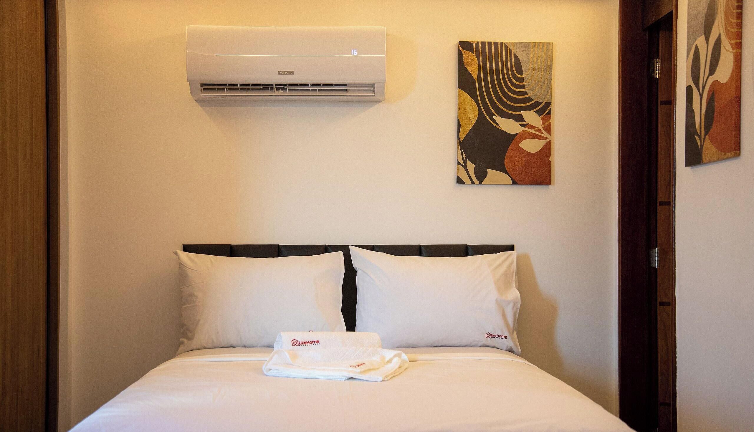 Habitación de hotel con una cama, almohadas blancas, una toalla doblada, un equipo de aire acondicionado y una obra de arte en la pared.