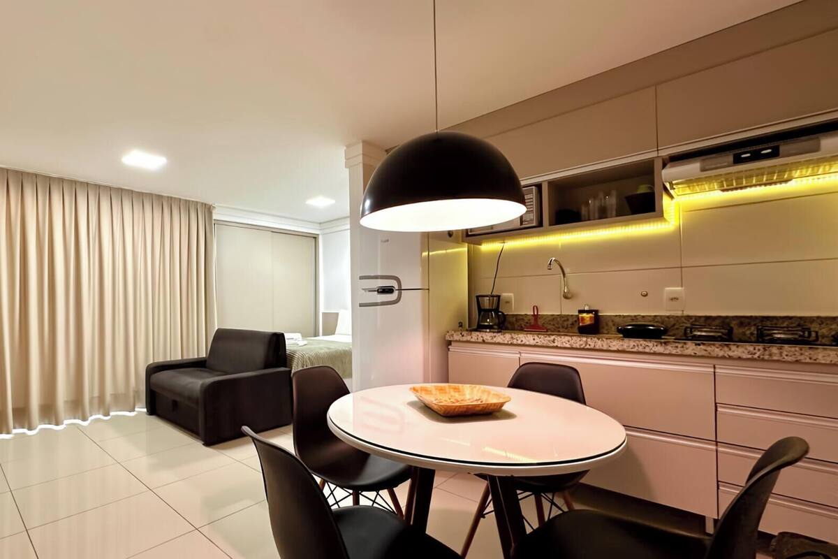 A modern hotel room with a dining area, a sofa, a TV, and a kitchen with a microwave and stove.