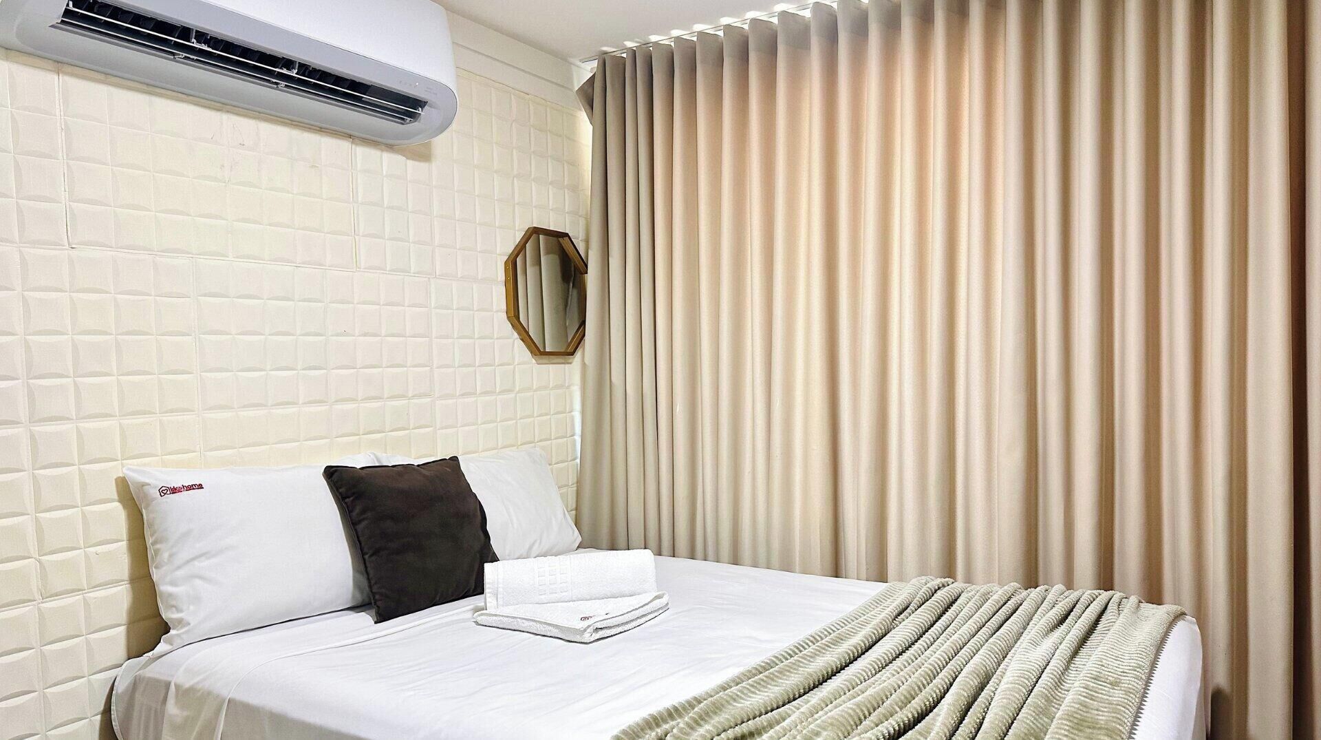 Habitación de hotel con cama, almohada blanca con logotipo, almohada oscura, toalla doblada, equipo de aire acondicionado y ventana con persianas verticales.