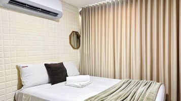 Quarto de hotel com uma cama, um travesseiro branco com um logotipo, um travesseiro escuro, uma toalha dobrada, um aparelho de ar condicionado e uma janela com venezianas verticais.