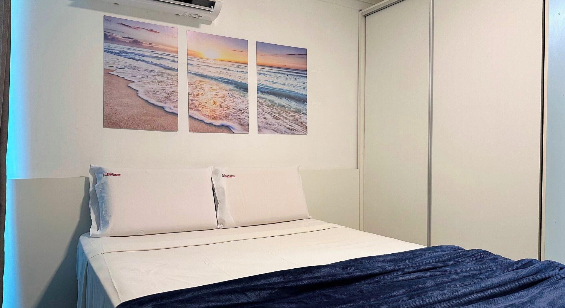 Una cama con ropa blanca de cama y una manta azul oscuro, un aire acondicionado instalado en la pared y tres cuadros con escenas de playa.