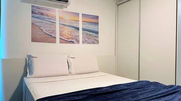 Una cama con ropa blanca de cama y una manta azul oscuro, un aire acondicionado instalado en la pared y tres cuadros con escenas de playa.
