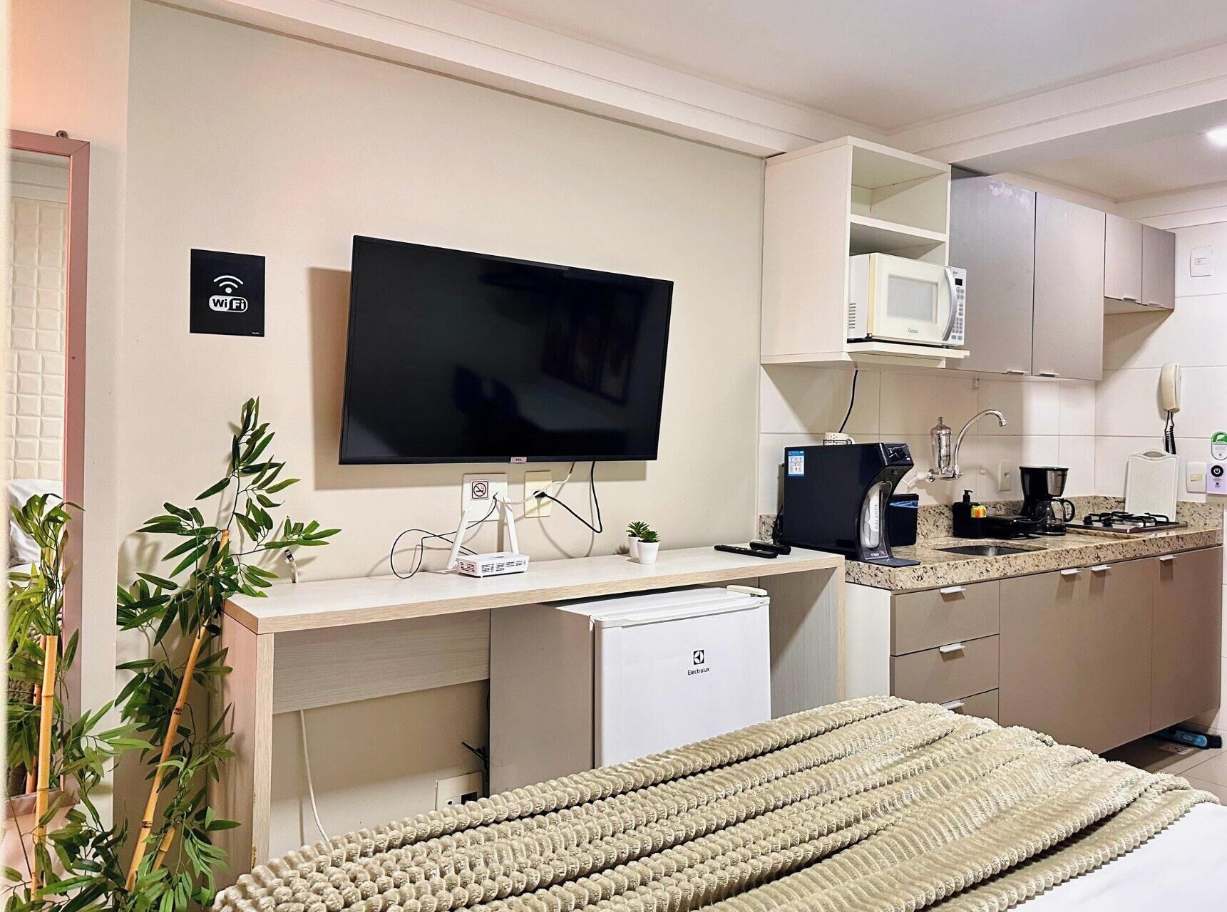 Quarto de hotel compacto com cozinha compacta, televisão de tela plana, pequeno frigorífico e cama com manta bege.