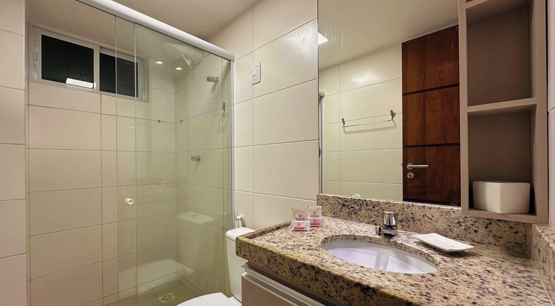 Un baño con ducha de vidrio, lavamanos de encimera de granito y un mueble de madera sobre el espejo.