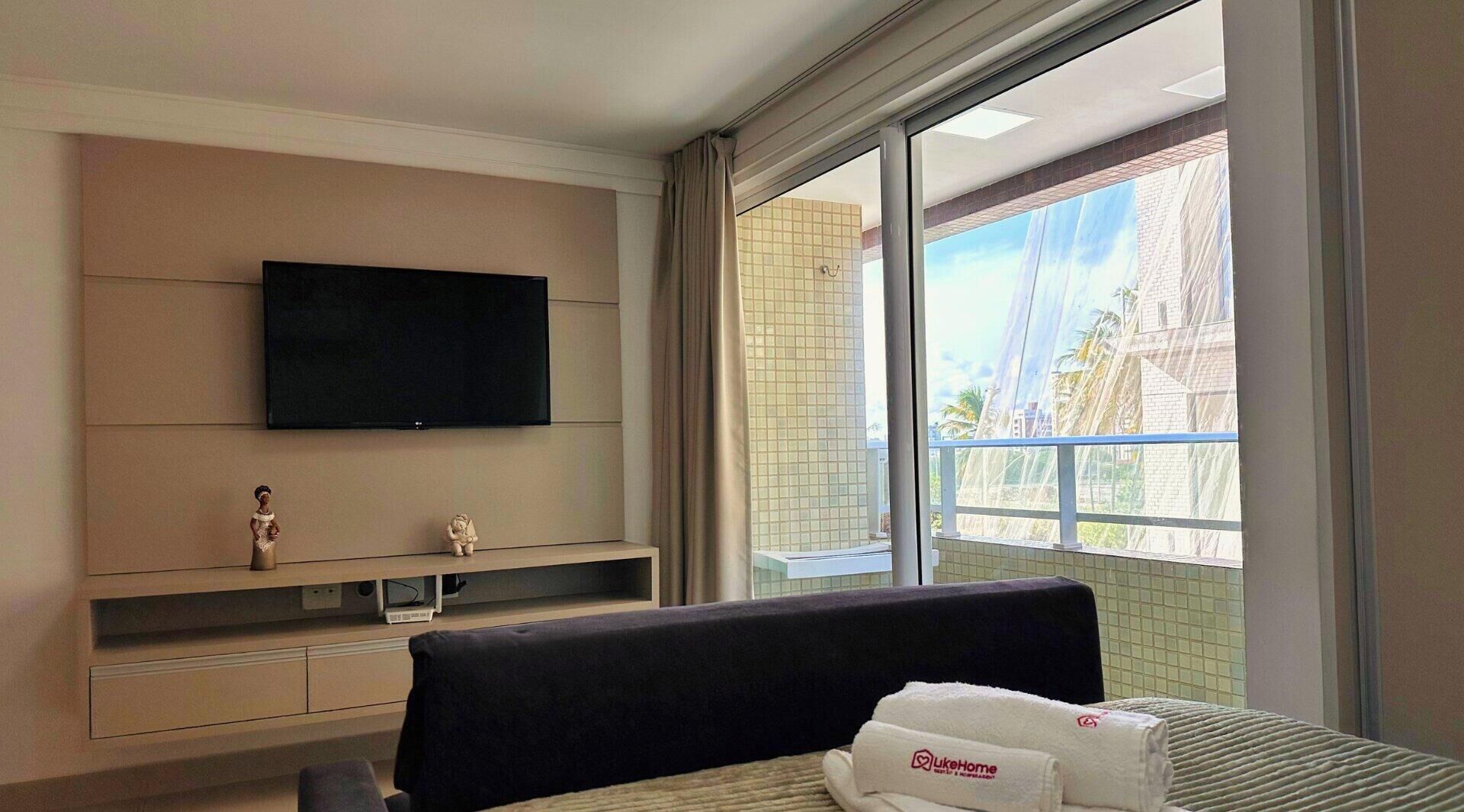 Habitació d'hotel amb televisió de pantalla plana penjada a la paret, llit amb capçalera, una petita estant amb figures i finestra amb vista a edificis i palmeres.
