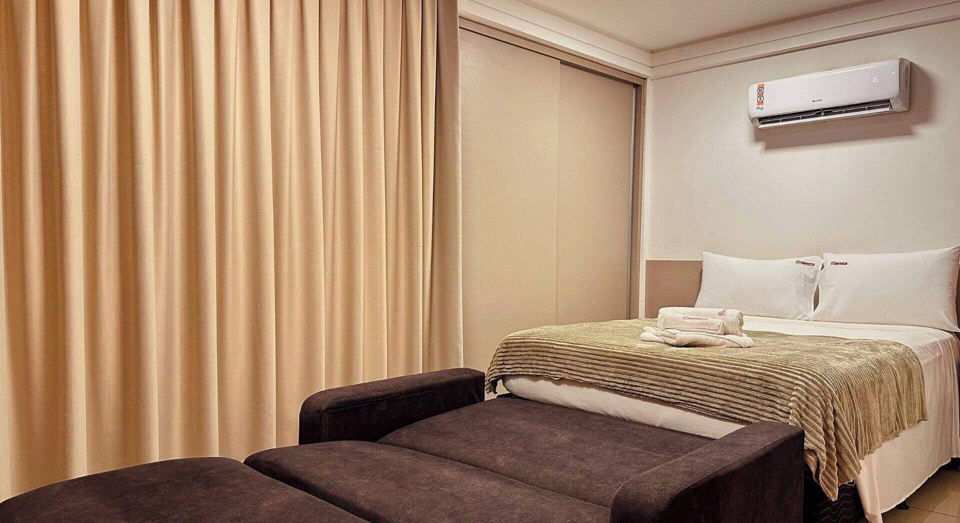 A hotel room with a bed, a chair, an air conditioning unit, and a curtain.