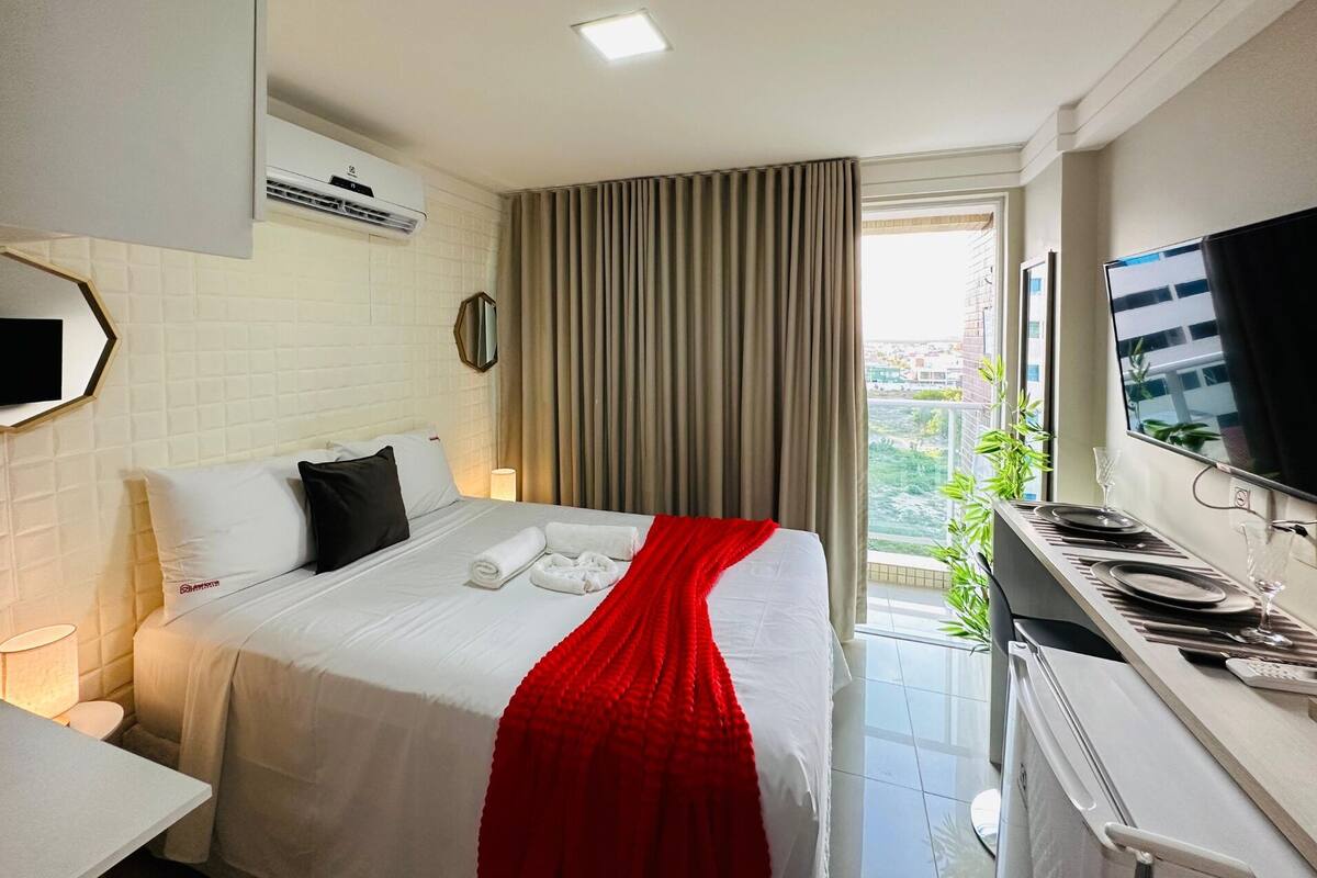 A hotel room with a bed, a TV, a desk with a computer, and a window with curtains.