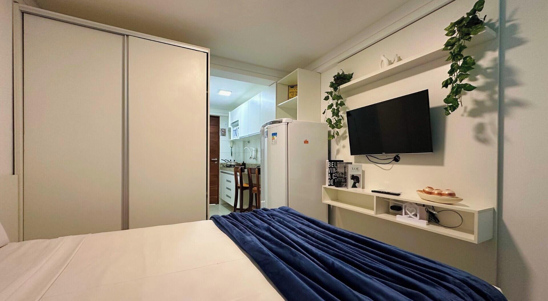 Un dormitorio con cama, televisor montado en la pared y una cocina visible a través de una puerta abierta.