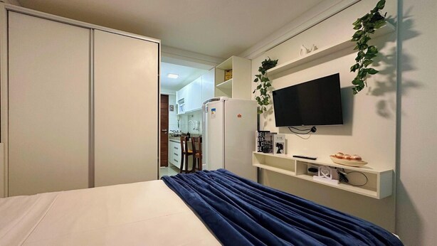 Un dormitorio con cama, televisor montado en la pared y una cocina visible a través de una puerta abierta.