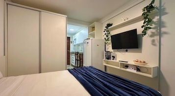 Un dormitorio con cama, televisor montado en la pared y una cocina visible a través de una puerta abierta.