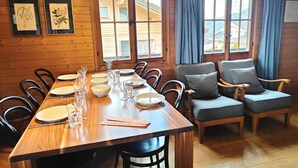 A wooden dining area with a long table set for six, surrounded by chairs. There are framed botanical prints on the wall, and large windows with curtains.
