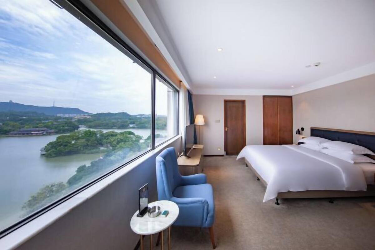 Habitación de hotel con una ventana grande, una cama, una silla, una mesita pequeña y una mesita de noche.