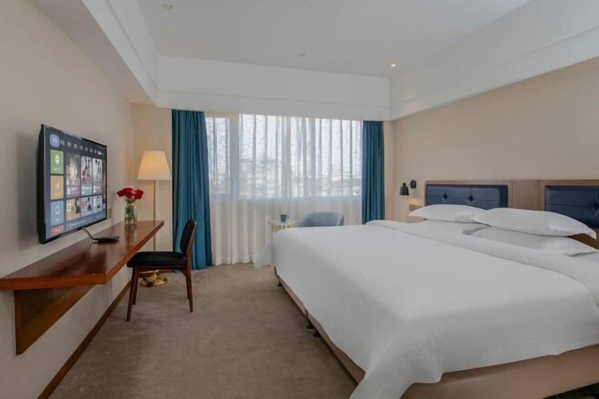Habitación de hotel con cama, escritorio con televisor, silla, lámpara y ventana con cortinas.
