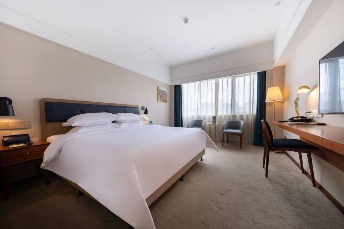 Habitación de hotel con una cama grande, un escritorio, una silla, una mesita de noche y una ventana con cortinas.