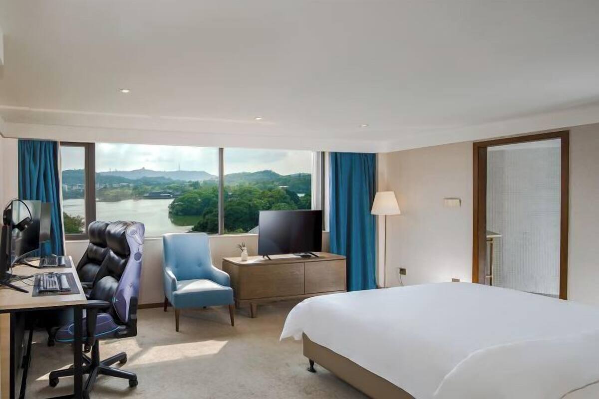 Habitación de hotel con cama, escritorio, ordenador, silla, televisor y vistas a un río y colinas.